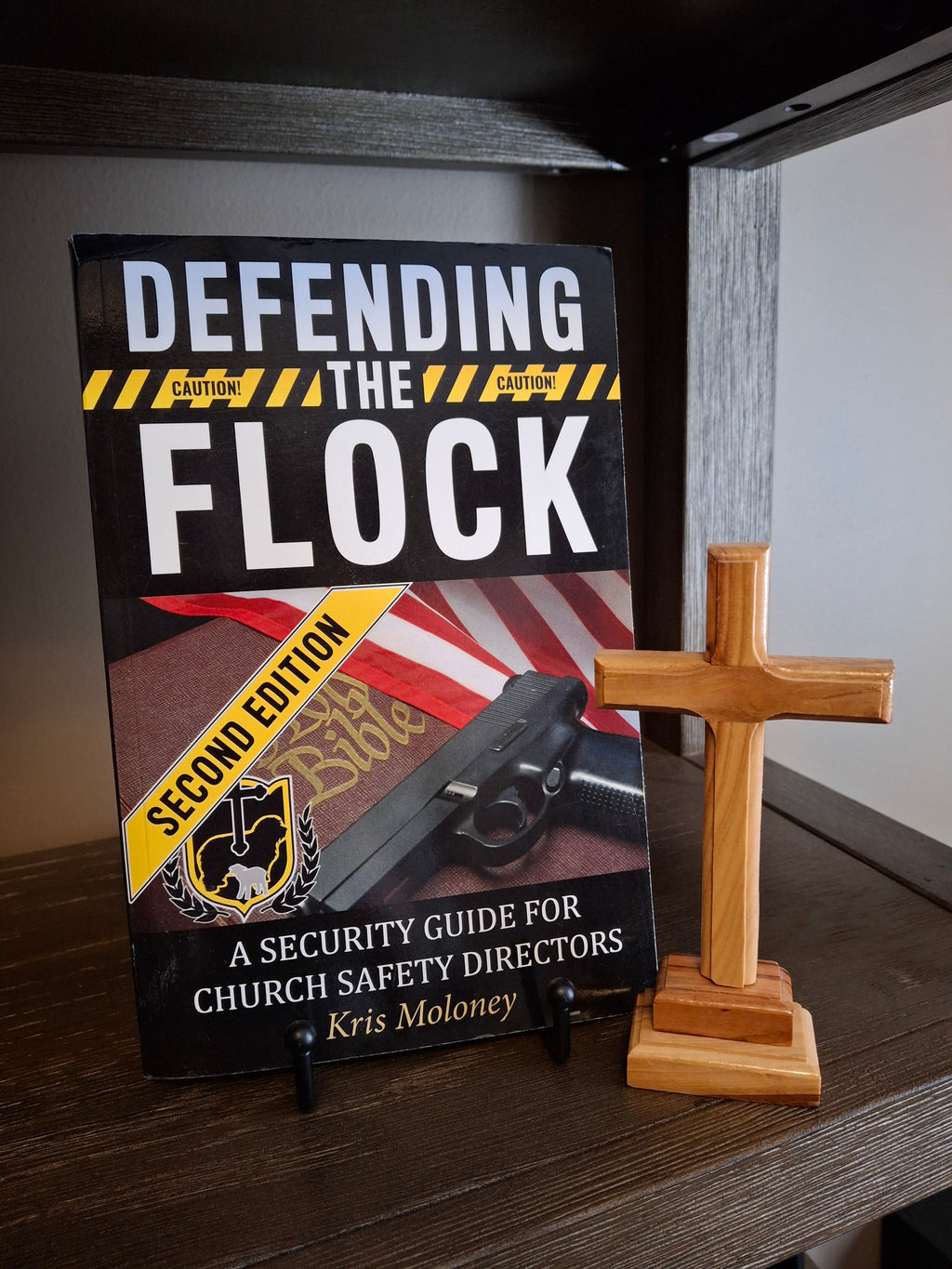 Defending the Flock paperback church security training manual for church safety directors displayed on a shelf next to a wooden cross