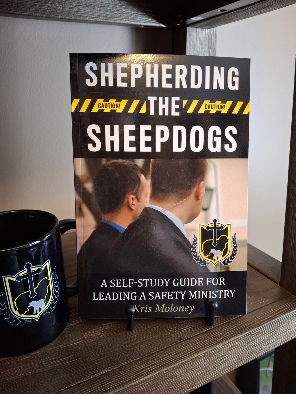 Shepherding the Sheepdogs paperback self-study guide for leading a church safety ministry displayed on a shelf next to an SCSA logo mug